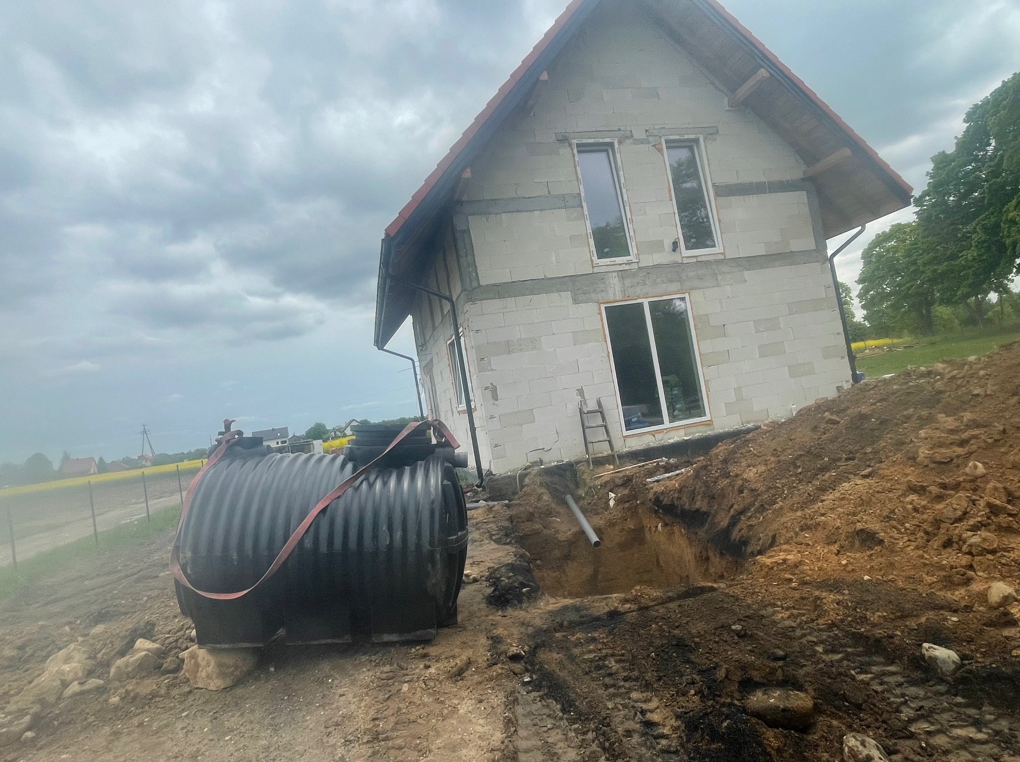Czarny zbiornik na szambo z czerwonym pasem transportowym, umieszczony obok wykopu, z widoczną rurą kanalizacyjną, na tle domu w stanie surowym i pochmurnego nieba.