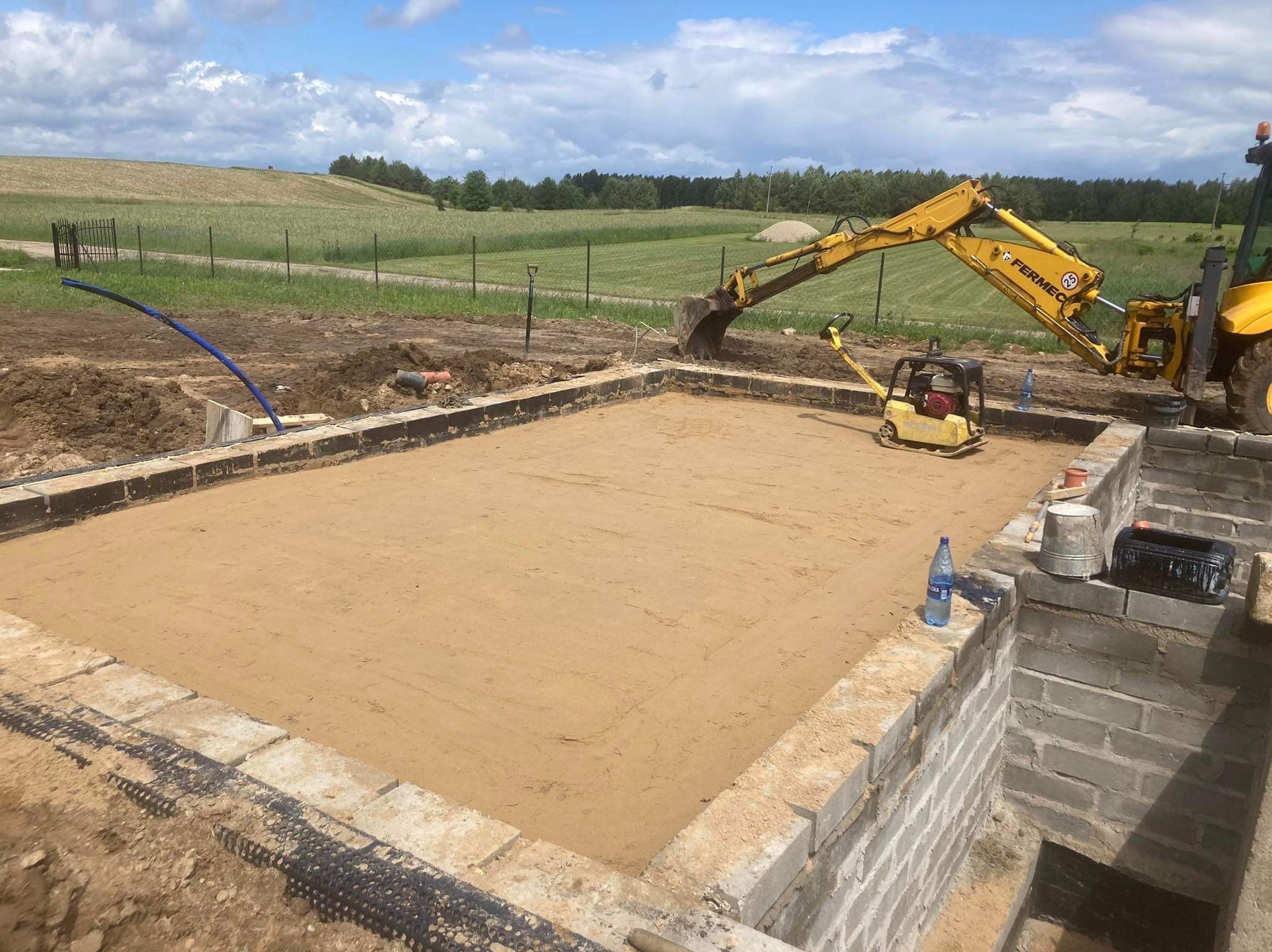 Wykop pod budynek z przygotowanym zbrojeniem, ubijarką, koparką Fermec w tle i widokiem na pola uprawne, słoneczny dzień.