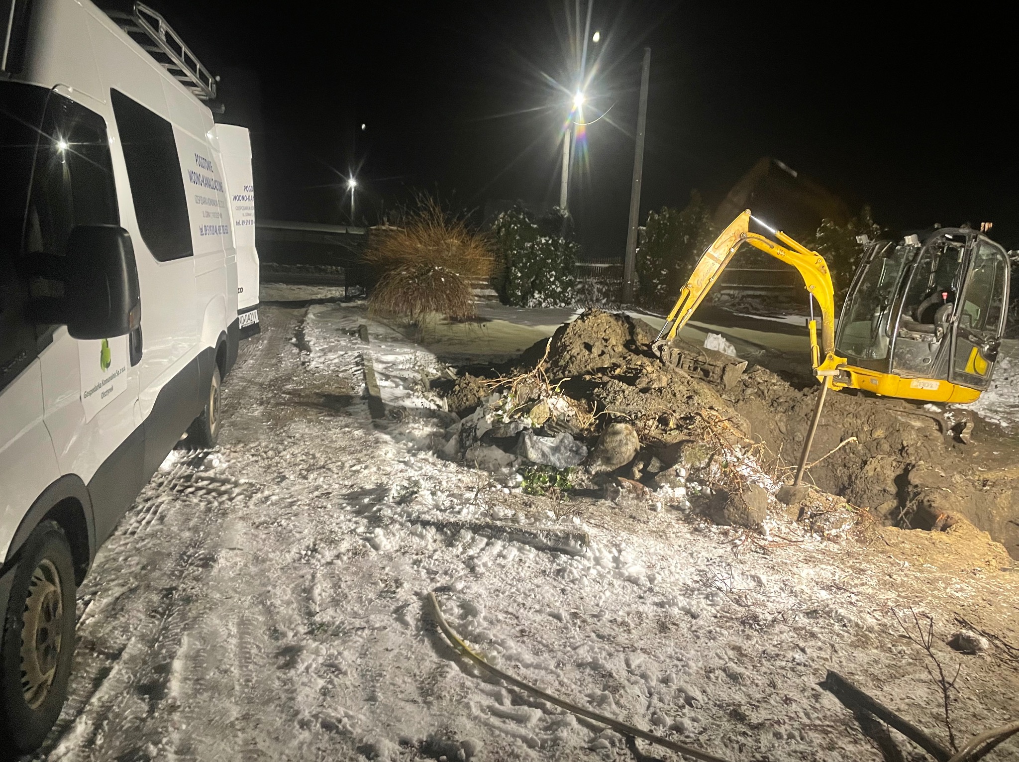 Żółta mini koparka wykonująca wykop w ziemi ośnieżonej śniegiem, nocne oświetlenie uliczne, biały samochód dostawczy firmy z widocznym logo i numerem telefonu w tle.
