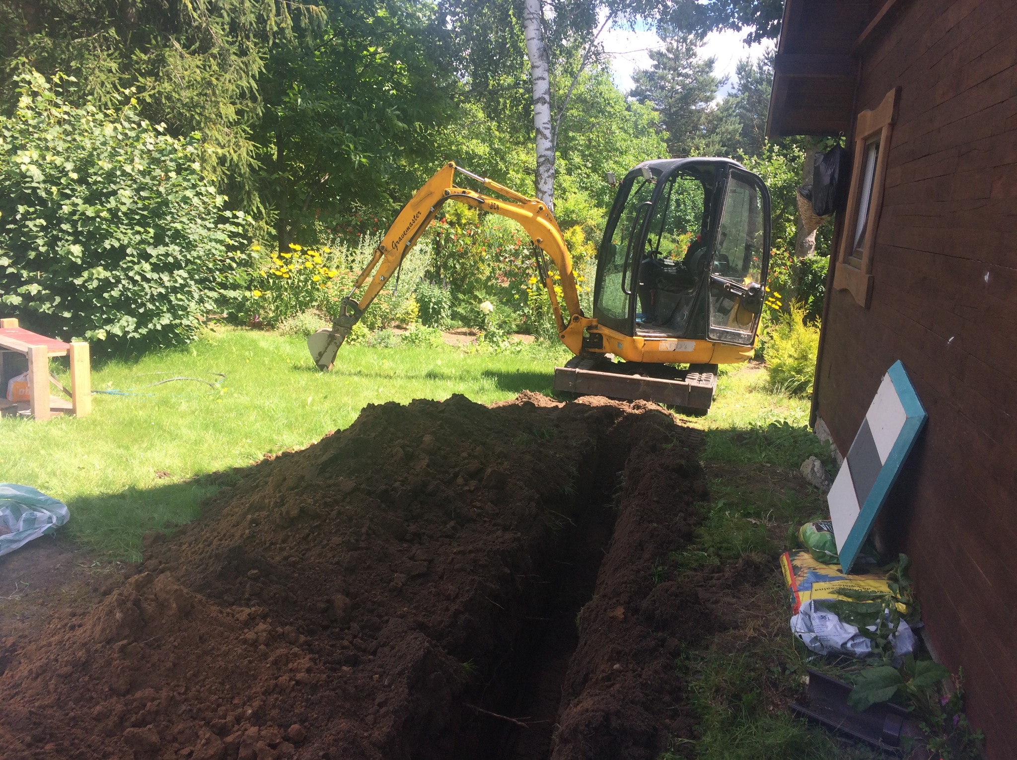 Żółta mini koparka Gravemaster obok wykopanego wąskiego rowu w trawiastym ogrodzie, przy drewnianym domku. Ziemia usypana wzdłuż rowu, obok domu leżą worki z ziemią.