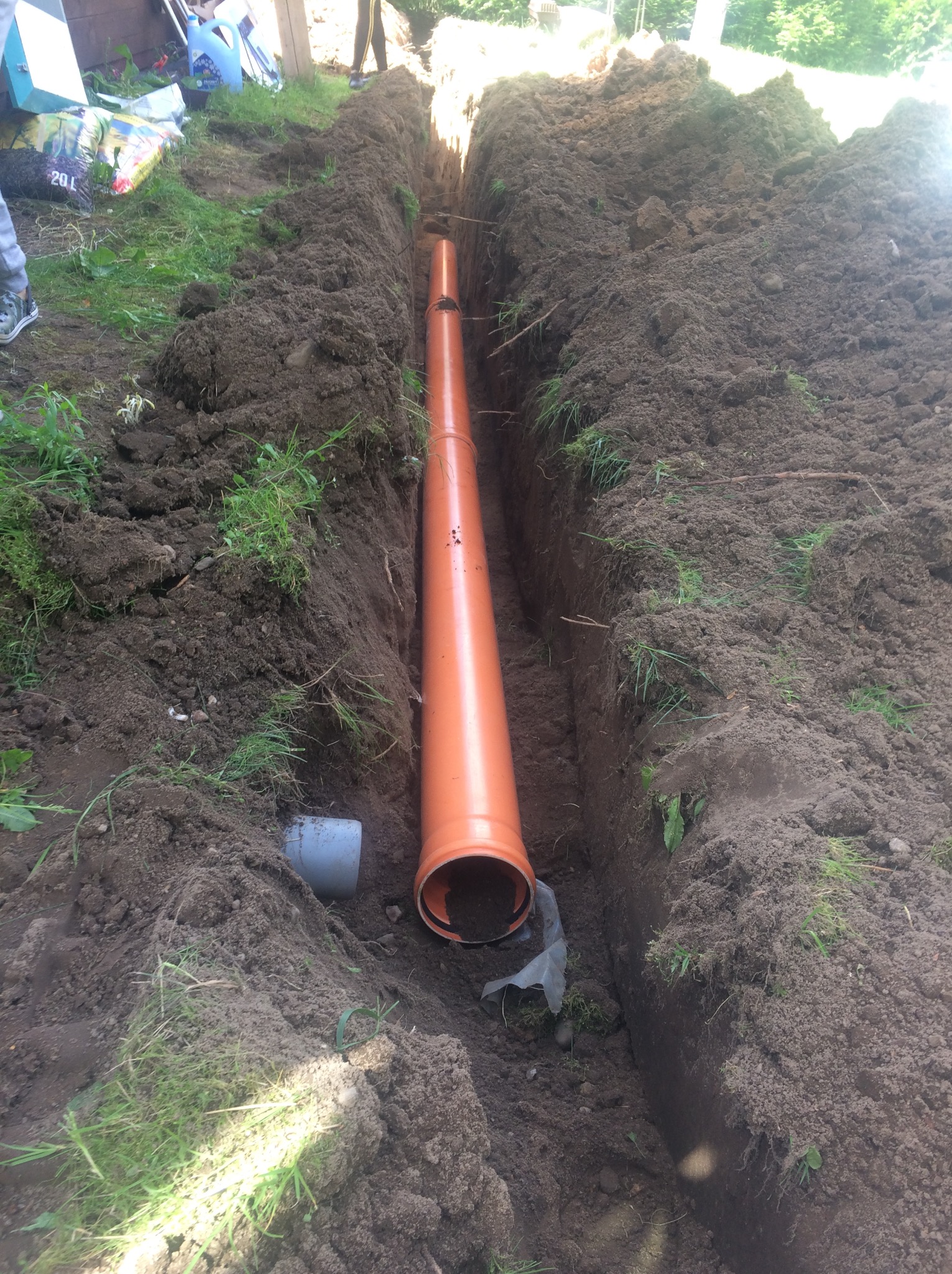 Pomarańczowa rura kanalizacyjna ułożona w wykopie ziemnym, widoczne ślady świeżo wykopanej ziemi i fragmenty trawy na brzegach wykopu.