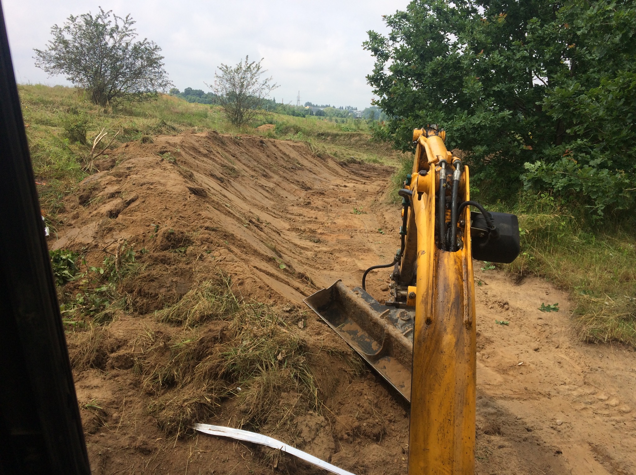 Żółta koparka stoi na skraju wykopu na nierównym terenie, z widocznymi śladami gąsienic, w tle drzewa i pochmurne niebo. Widok z wnętrza kabiny.