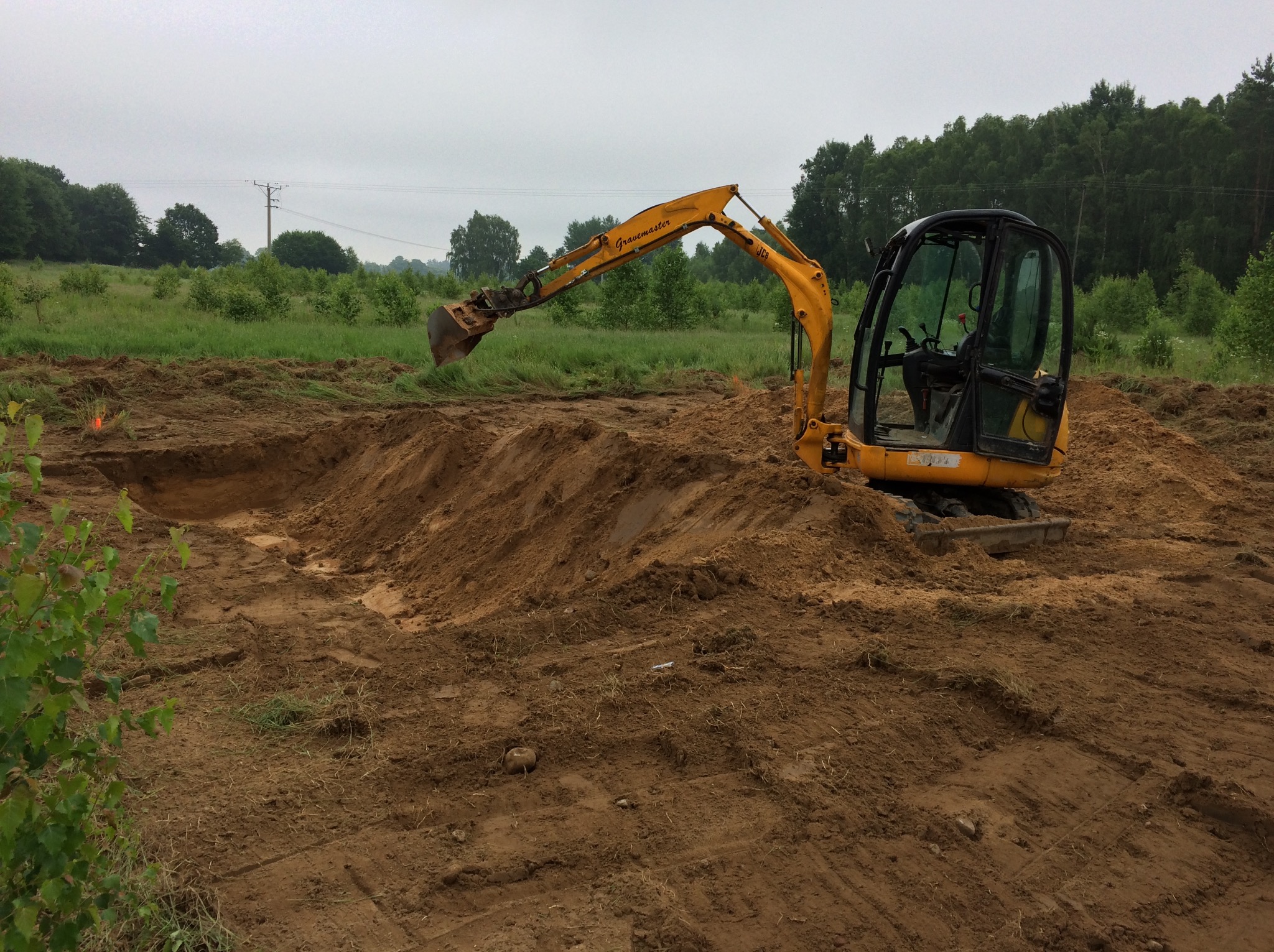 Żółta minikoparka JCB Gravemaster na placu budowy, w trakcie prac ziemnych, widoczne wykopane zagłębienie w ziemi, w tle zieleń i pochmurne niebo.