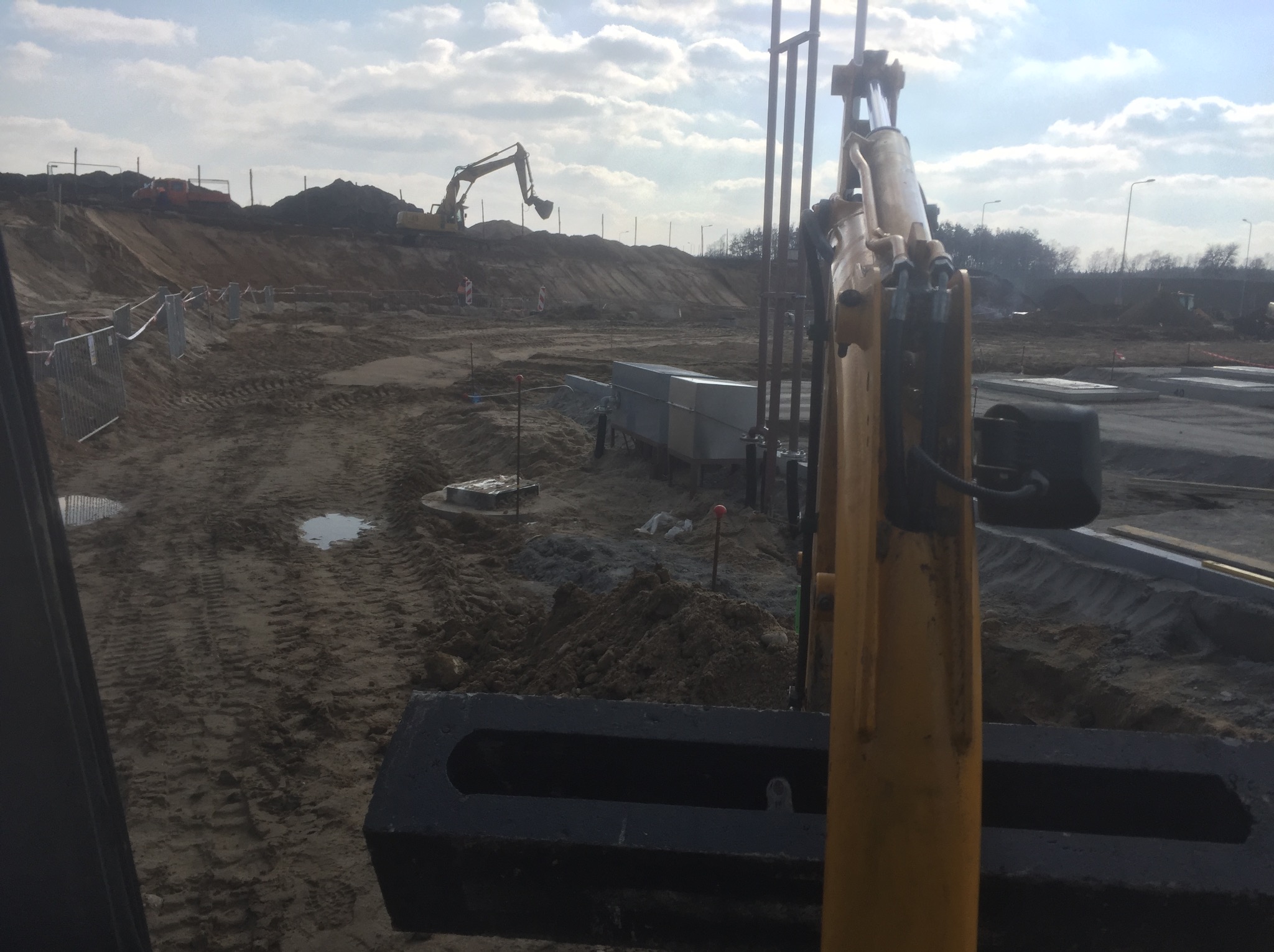 Żółty wysięgnik koparki na pierwszym planie, w tle plac budowy z koparką pracującą na usypanym wzgórzu ziemi, widoczne elementy konstrukcyjne i kałuże na grząskim terenie.