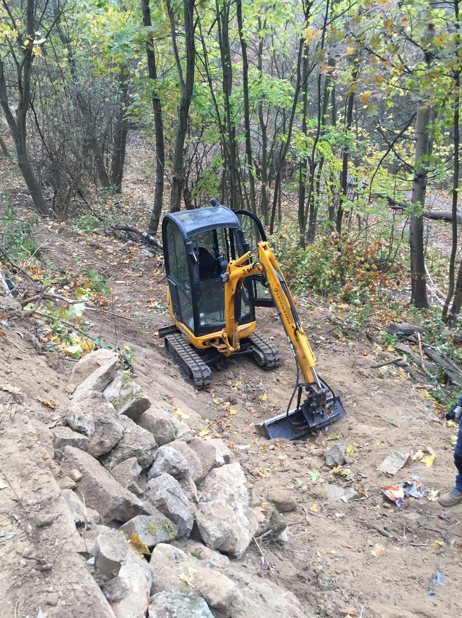 Żółta mini koparka na gąsienicach, z otwartą kabiną, stoi na zboczu ziemnym w lesie, obok sterty kamieni i śmieci, przygotowując teren pod budowę.