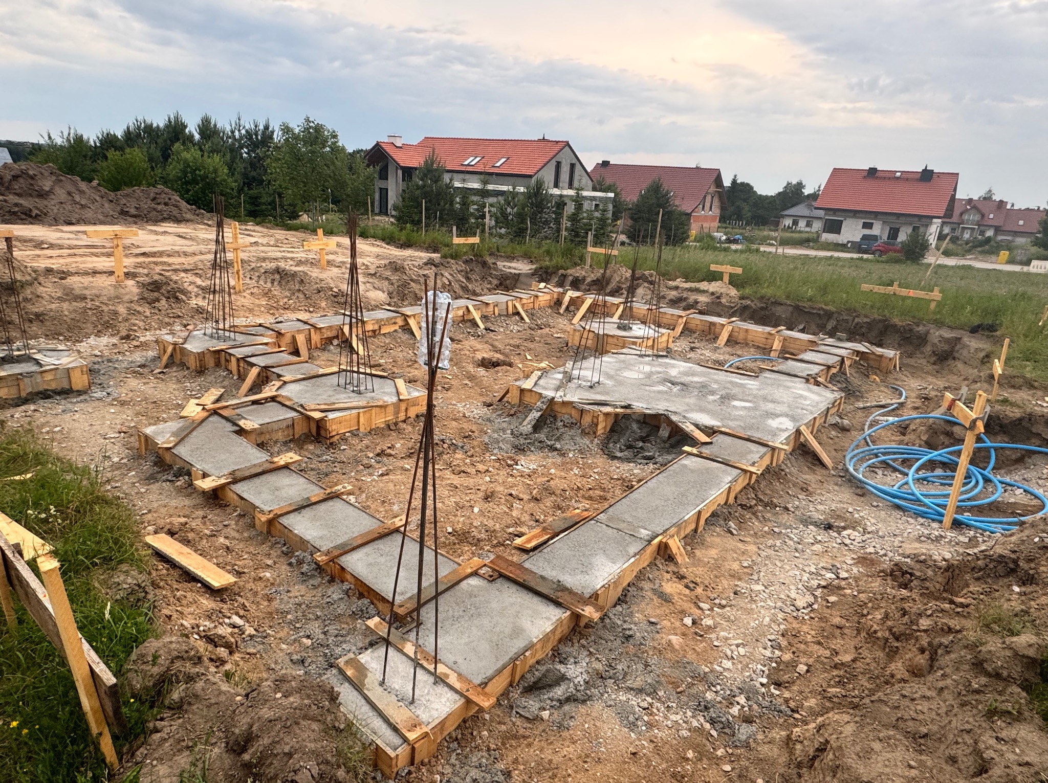 Zalane betonem szalunki pod fundamenty domu w Olsztynku. Widoczne wystające pręty zbrojeniowe i niebieski wąż. W tle zabudowa jednorodzinna i pochmurne niebo.