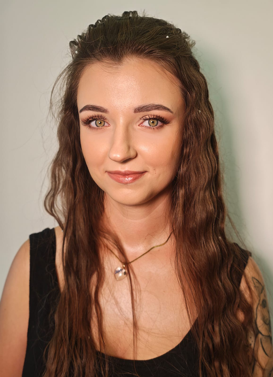 Portret młodej kobiety z profesjonalnym makijażem, podkreślone oczy i usta, delikatne upięcie włosów z ozdobnymi wstawkami, widoczny tatuaż na ramieniu.