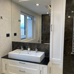 Gorzewski Meble - Nowoczesna łazienka z białą szafką pod umywalkę z czarnym blatem, dużą umywalką nablatową i wysoką białą szafką, odbicie okna w dużym lustrze.