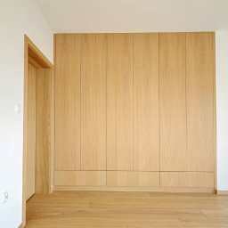 Gorzewski Meble - Wnętrze z dużą, drewnianą szafą wnękową z jasnego drewna, dopasowaną kolorystycznie do podłogi, z widocznym fragmentem ościeżnicy drzwiowej z tego samego materiału.