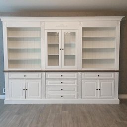 Gorzewski Meble - Biała, drewniana biblioteka na wymiar z przeszklonymi drzwiczkami na środku i szufladami na dole, z ciemnym blatem i dekoracyjnymi listwami.