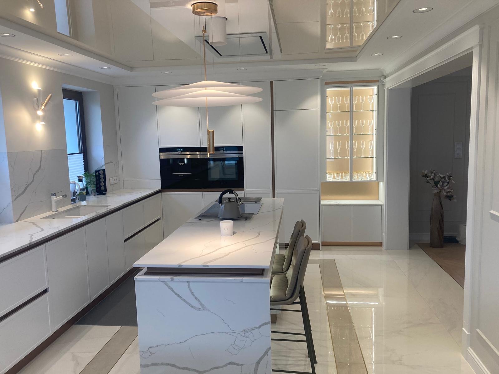 Jasna kuchnia na wymiar z wyspą i blatem imitującym marmur, oświetlona nowoczesną lampą wiszącą i kinkietem, z przeszkloną witryną na kieliszki w tle.