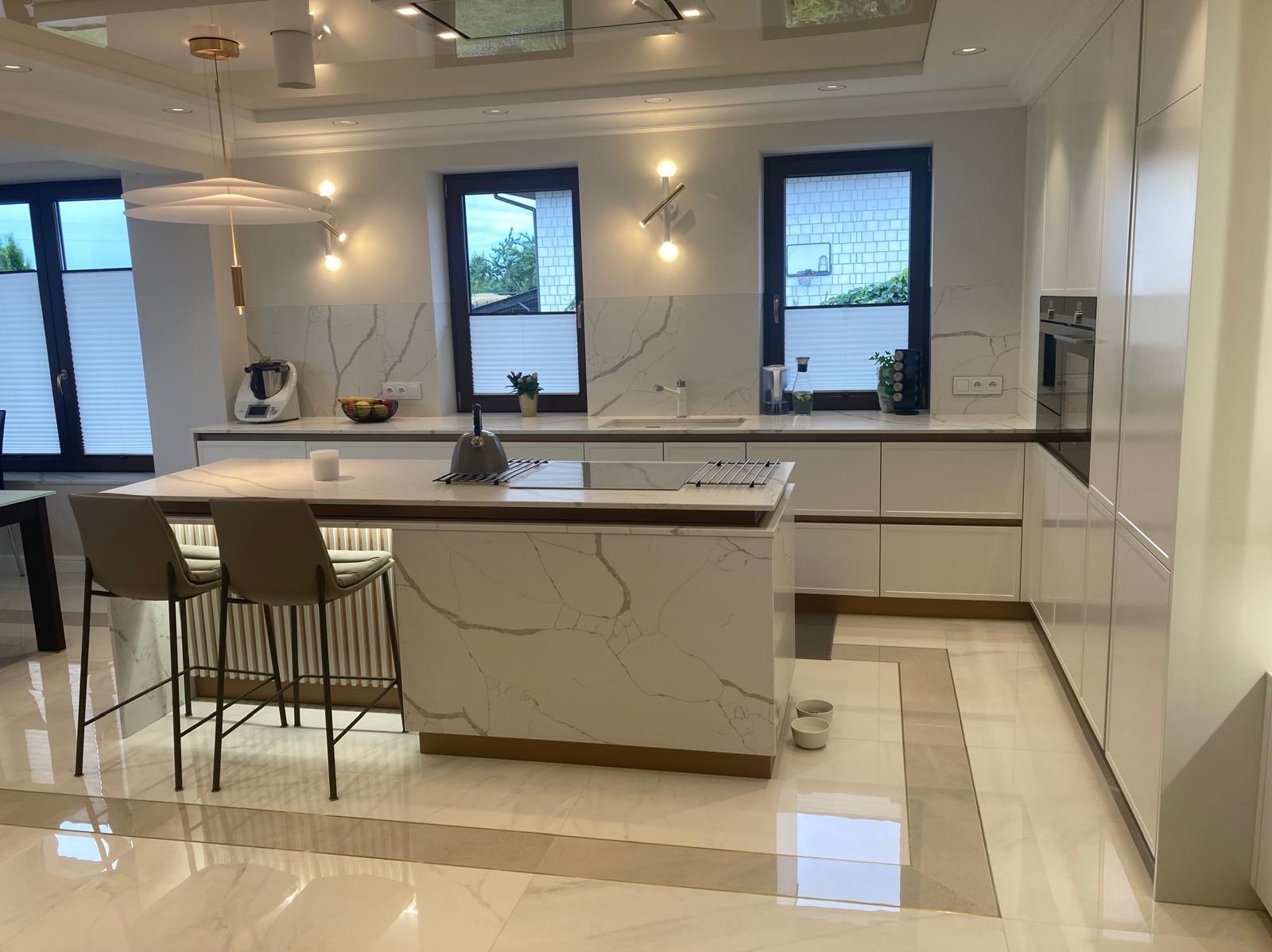 Nowoczesna kuchnia z wyspą wykończoną białym marmurem, oświetlona lampą wiszącą, z dwoma hokerami przy wyspie, widok na okna z roletami plisowanymi.