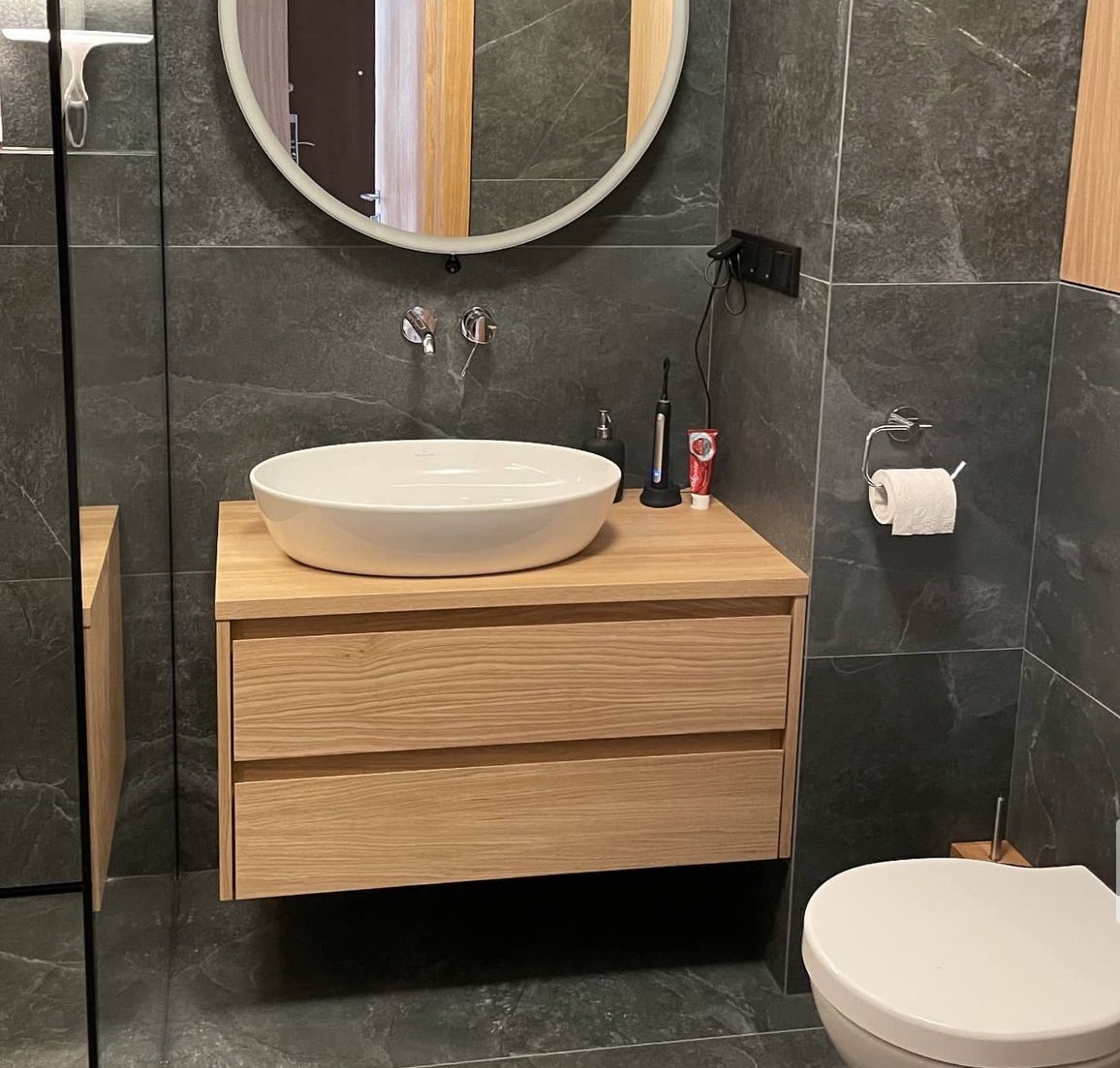 Nowoczesna łazienka z umywalką nablatową na szafce z jasnego drewna, okrągłe lustro z podświetleniem, szare płytki imitujące kamień na ścianach i podłodze, widoczna szczoteczka elektryczna...