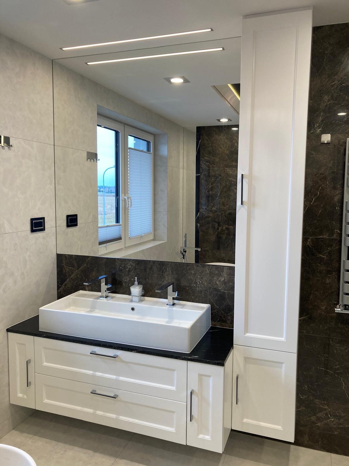 Nowoczesna łazienka z białą szafką pod umywalkę z czarnym blatem, dużą umywalką nablatową i wysoką białą szafką, odbicie okna w dużym lustrze.