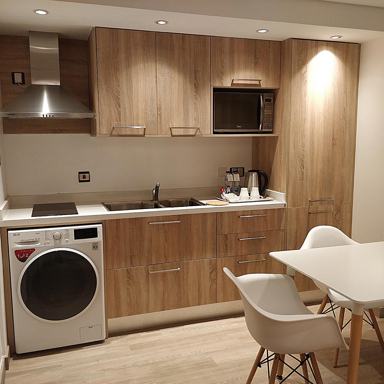Nowoczesna kuchnia z drewnianymi frontami, zintegrowana z pralką, minimalistyczna aranżacja z białym stołem i krzesłami, oświetlenie punktowe.