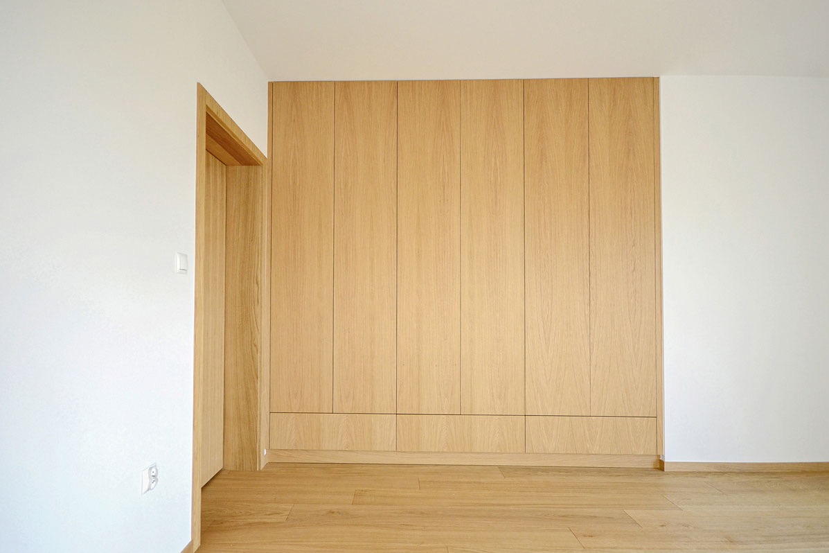 Wnętrze z dużą, drewnianą szafą wnękową z jasnego drewna, dopasowaną kolorystycznie do podłogi, z widocznym fragmentem ościeżnicy drzwiowej z tego samego materiału.