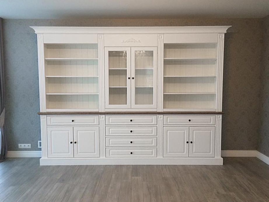 Biała, drewniana biblioteka na wymiar z przeszklonymi drzwiczkami na środku i szufladami na dole, z ciemnym blatem i dekoracyjnymi listwami.