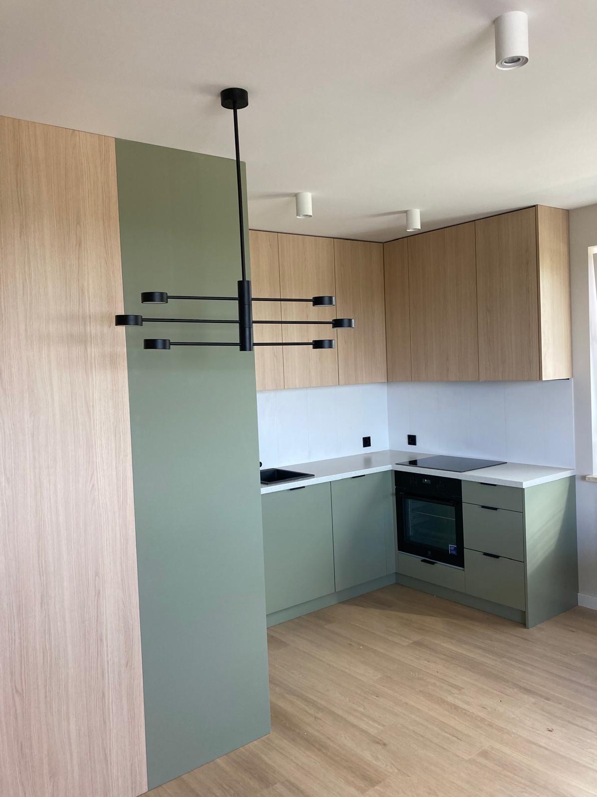Nowoczesna kuchnia z frontami w odcieniach zieleni i drewna, minimalistyczny czarny żyrandol, biały blat i płytki, drewniana podłoga, widok na narożną zabudowę z piekarnikiem.