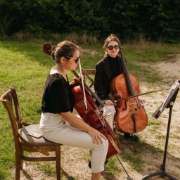 Dolcello - Oprawa Muzyczna - Dwie kobiety w okularach siedzą na krzesłach na trawie, grając na wiolonczelach, z nutami na stojaku obok. Jedna z kobiet ma telefon leżący na krześle obok niej.