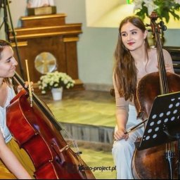Dolcello - Oprawa Muzyczna - Dwie młode kobiety grające na wiolonczelach w kościele, jedna w żółtej spódnicy patrzy na drugą, która uśmiecha się patrząc na nuty.
