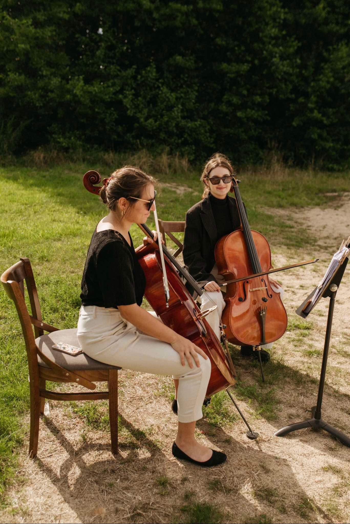Dwie kobiety w okularach siedzą na krzesłach na trawie, grając na wiolonczelach, z nutami na stojaku obok. Jedna z kobiet ma telefon leżący na krześle obok niej.
