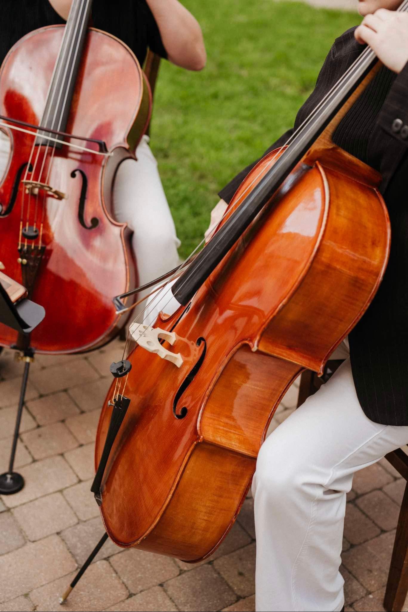 Dwaj mężczyźni siedzą na krzesłach, trzymając wiolonczele na tle zielonego trawnika i kostki brukowej.