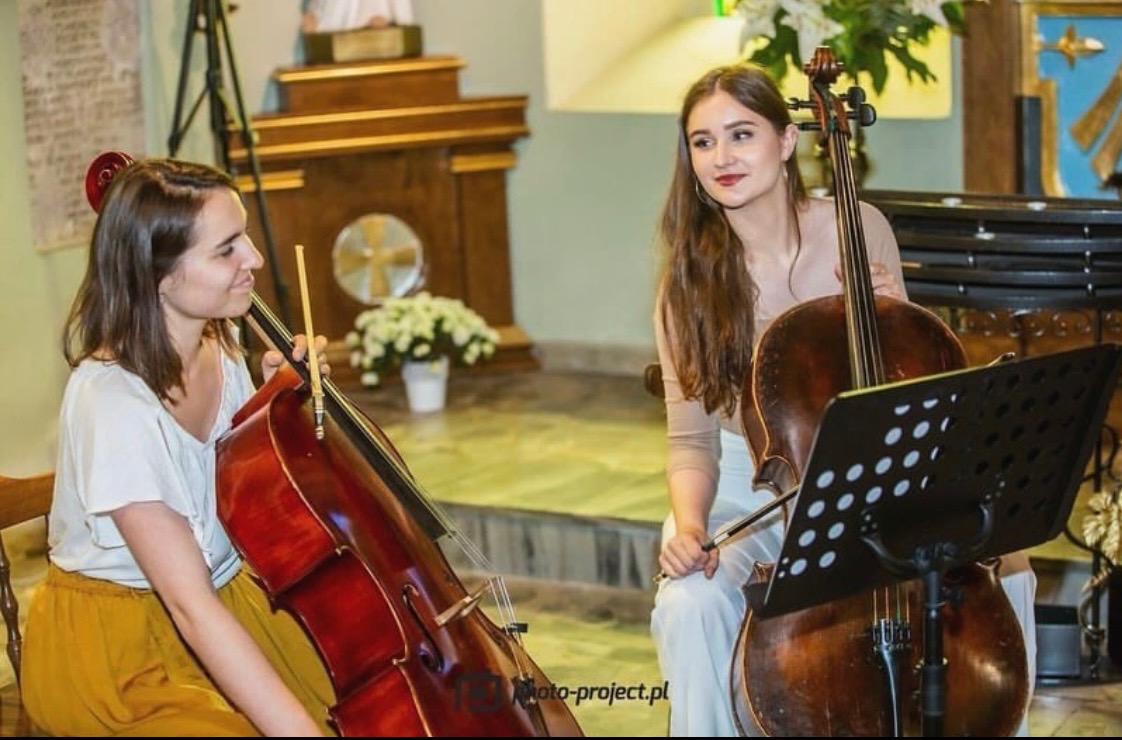 Dwie młode kobiety grające na wiolonczelach w kościele, jedna w żółtej spódnicy patrzy na drugą, która uśmiecha się patrząc na nuty.