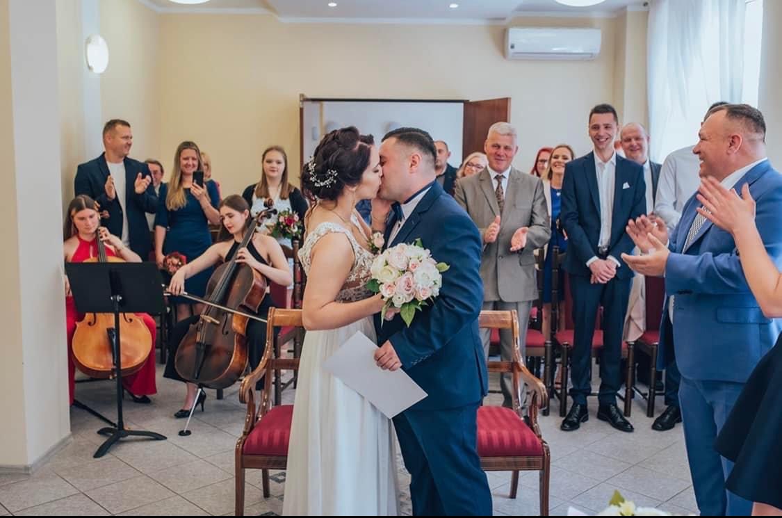 Para młoda całuje się podczas ceremonii ślubnej, w tle zespół smyczkowy gra na wiolonczelach i skrzypcach, a goście klaszczą.