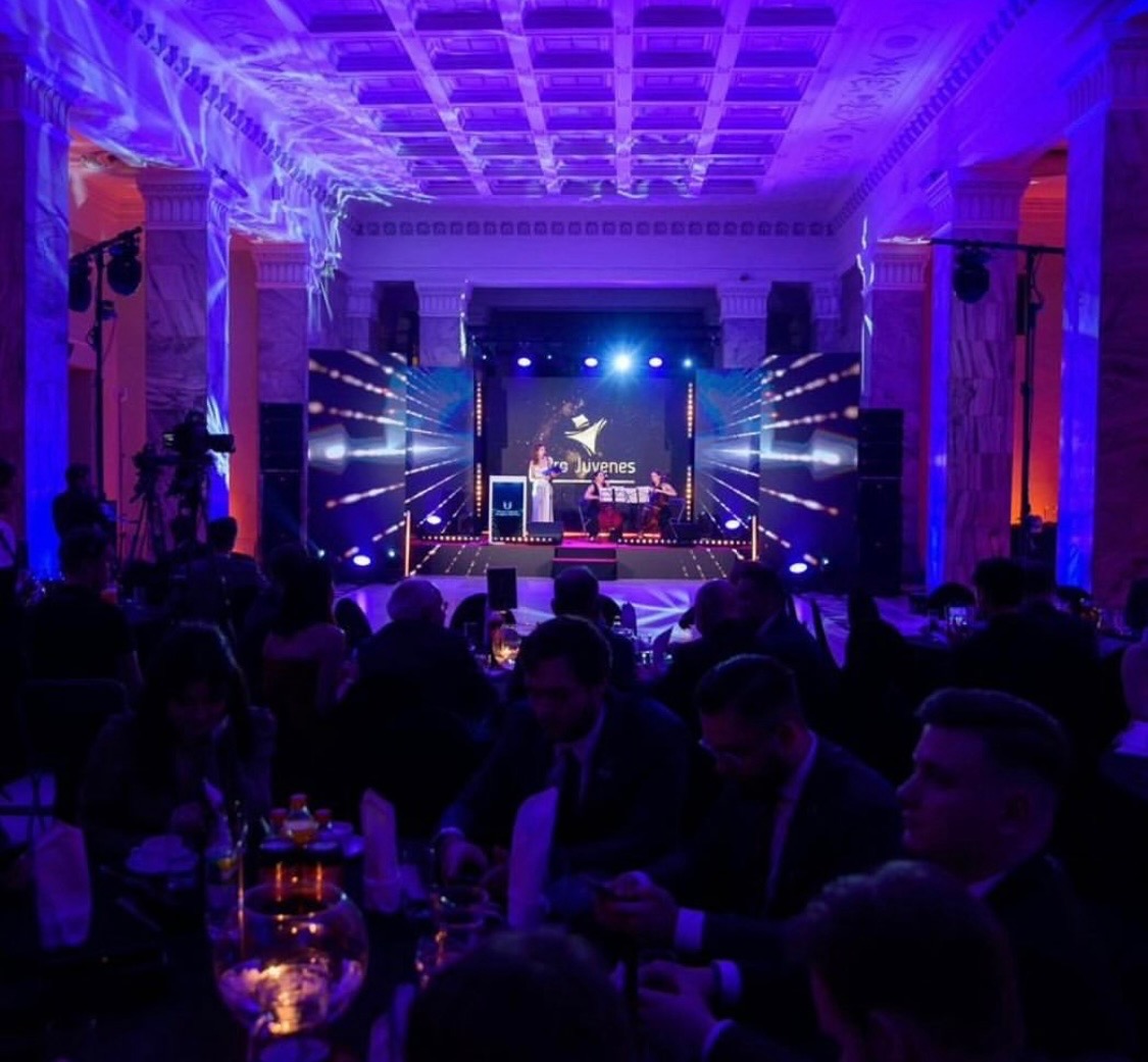 Sala balowa z widokiem na scenę podczas występu zespołu muzycznego i wokalistki, oświetlenie sceniczne w odcieniach fioletu i niebieskiego, publiczność przy stołach.