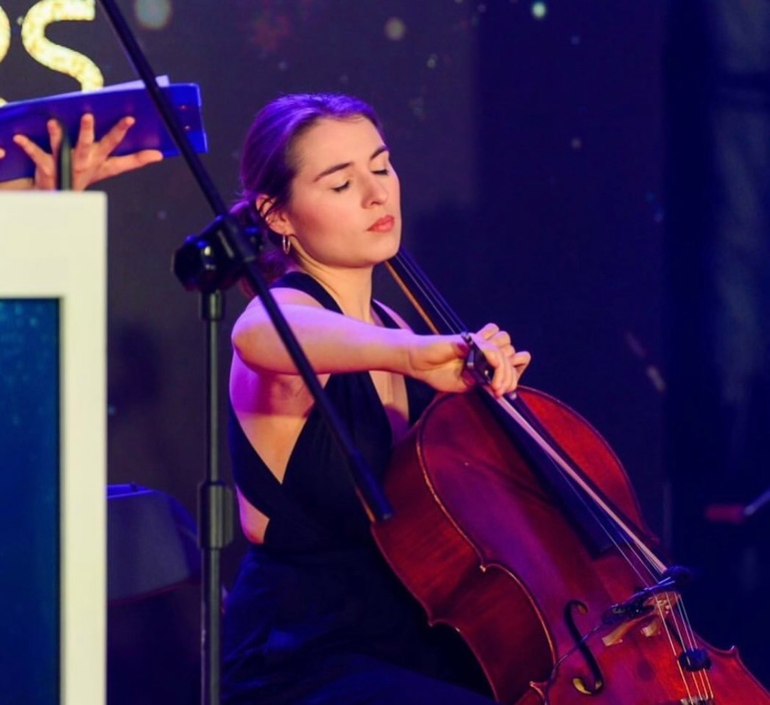 Młoda kobieta w czarnej sukni gra na wiolonczeli na scenie podczas występu, z zamkniętymi oczami i skupionym wyrazem twarzy.