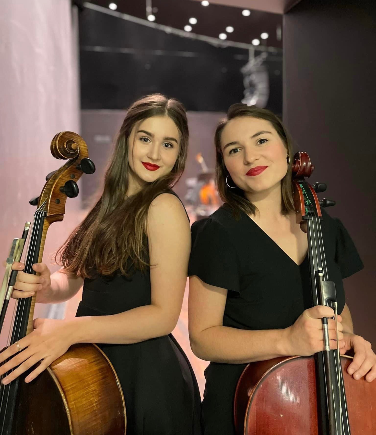 Dwie młode kobiety, ubrane w czarne sukienki, pozują z wiolonczelami na tle sceny z widocznymi instrumentami w tle.