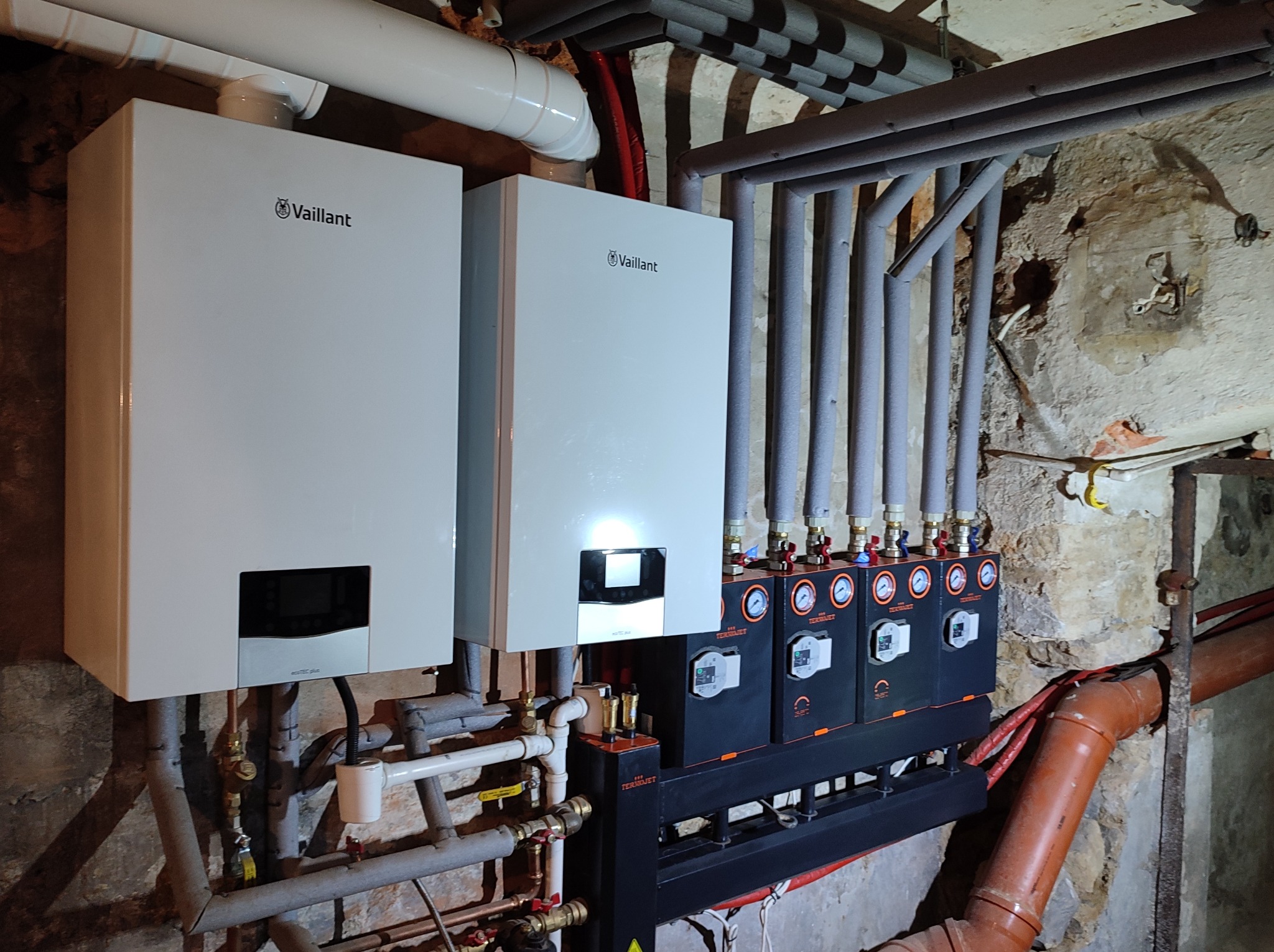 Dwa białe kotły gazowe Vaillant zamontowane w piwnicy z widocznymi rurami, zaworami, czujnikami i instalacją w surowym stanie.