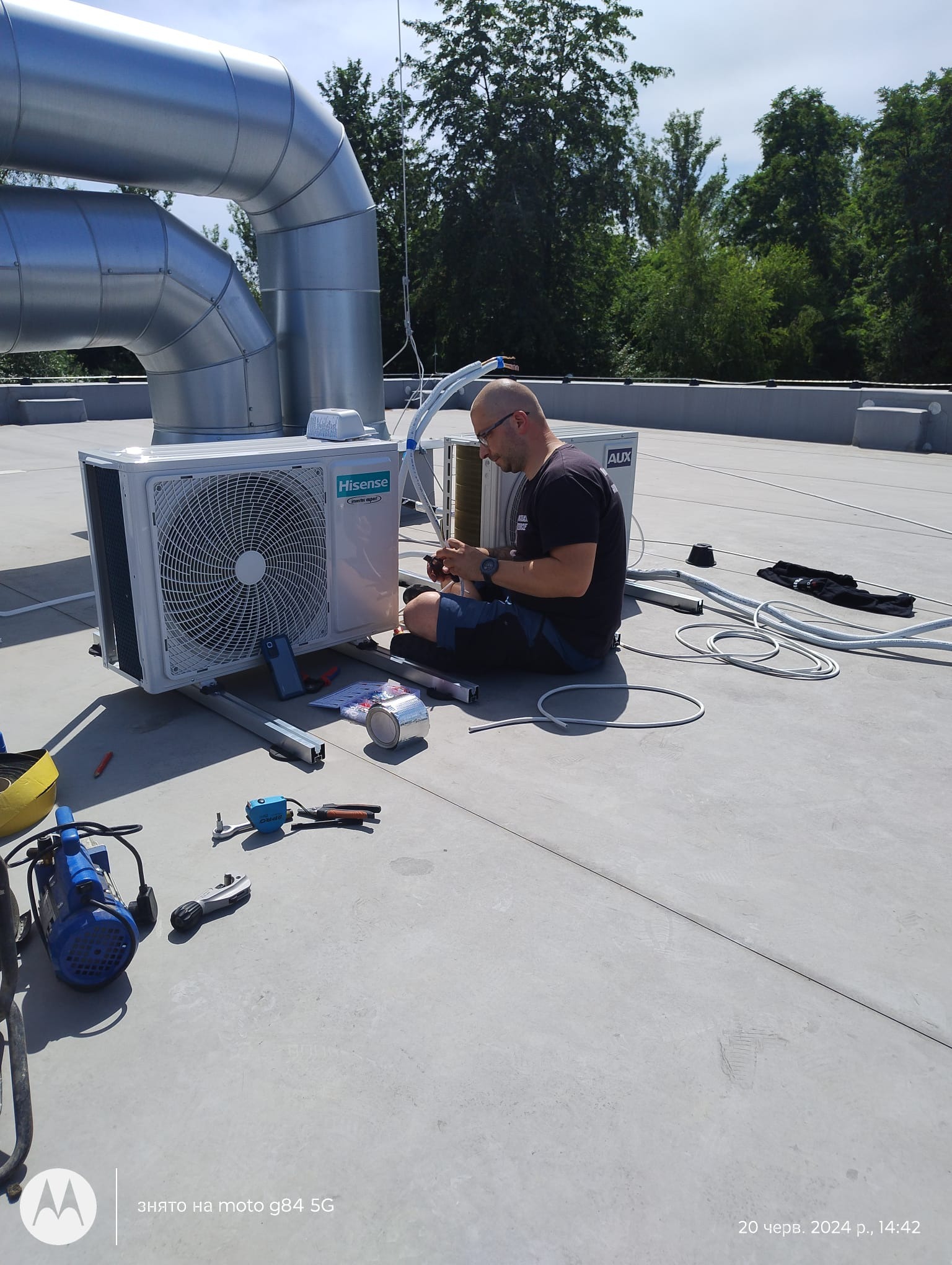 Instalator siedzi na dachu obok jednostki zewnętrznej klimatyzacji Hisense i AUX, podłączając przewody. Widoczne narzędzia, rury wentylacyjne i drzewa w tle.
