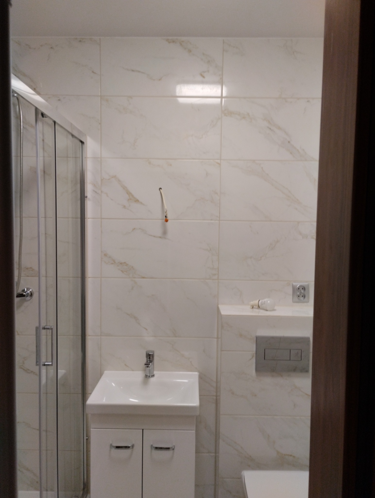 Wykończona łazienka z kabiną prysznicową, umywalką z szafką, toaletą i płytkami imitującymi marmur. Widoczny niedokończony montaż oświetlenia.