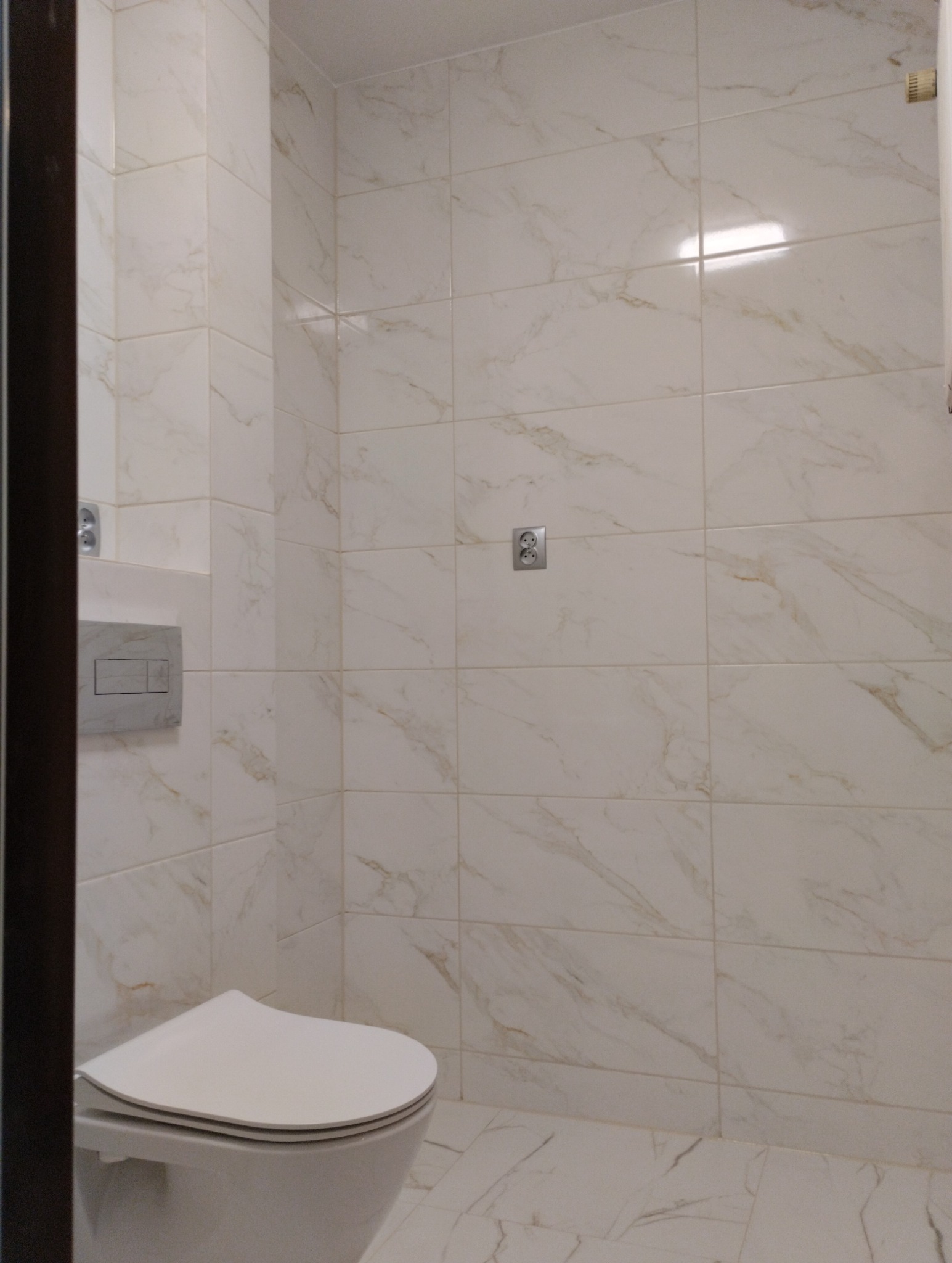 Biała toaleta z podwieszanym zbiornikiem i przyciskiem spłukującym na ścianie wyłożonej płytkami imitującymi marmur, widoczne gniazdko elektryczne.