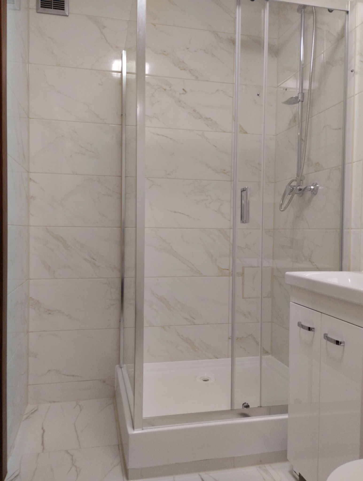 Nowoczesna kabina prysznicowa z przezroczystego szkła i chromowanymi detalami, zamontowana w łazience wykończonej płytkami imitującymi marmur, obok biała szafka pod umywalkę z chromowanymi uchwytami.