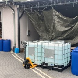 Skład Opału Klikawa TJ MOBILE Sp. z o.o. - Magazyn z paletami brykietu drzewnego w tle, beczkami i kontenerami IBC z płynem, oraz wózkiem paletowym na kostce brukowej przed budynkiem.