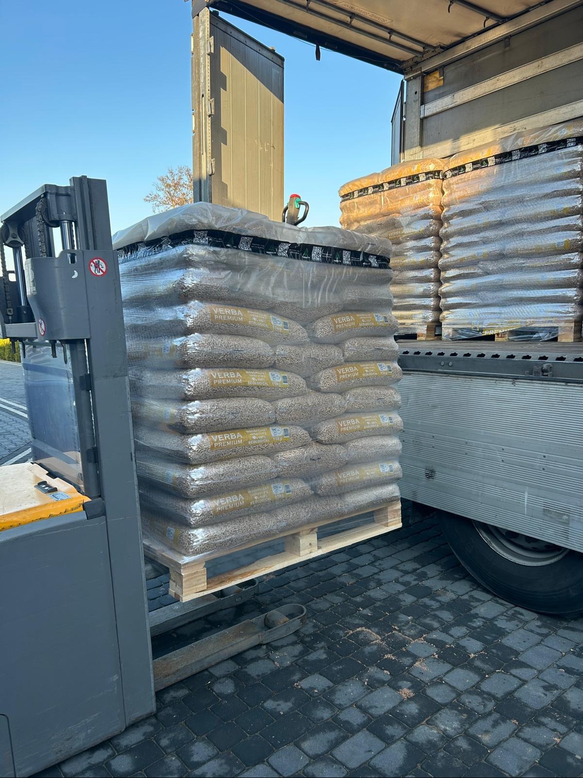 Wózek widłowy podnosi paletę z workami pelletu marki Verba Premium, widoczna naczepa ciężarówki w trakcie rozładunku.