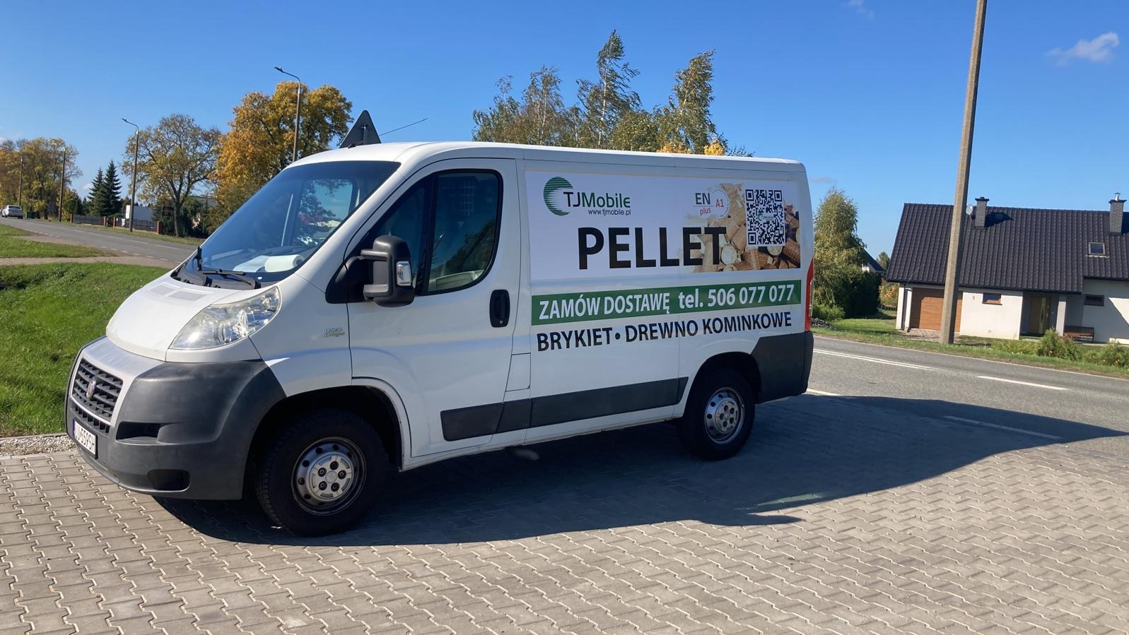 Biały samochód dostawczy z reklamą pelletu, brykietu i drewna kominkowego firmy TJMobile, zaparkowany na brukowanej powierzchni, z widocznym domem w tle.