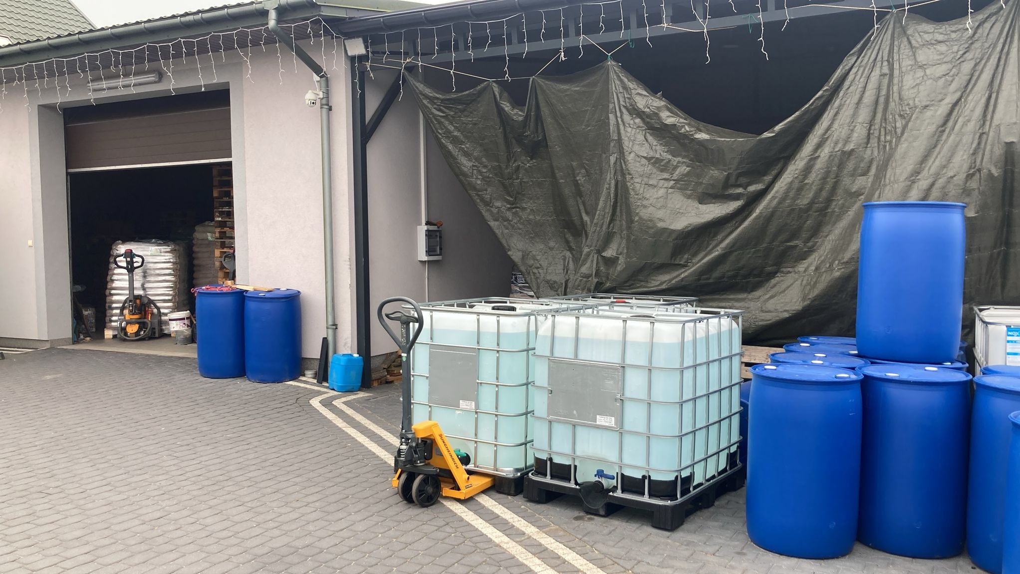 Magazyn z paletami brykietu drzewnego w tle, beczkami i kontenerami IBC z płynem, oraz wózkiem paletowym na kostce brukowej przed budynkiem.