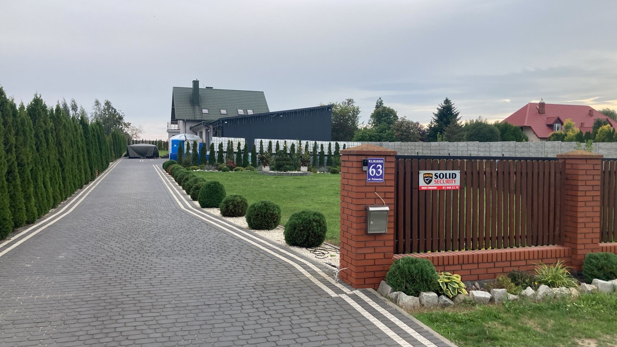 Podjazd z kostki brukowej prowadzący do domu z zadbanym ogrodem, ogrodzeniem z drewna i cegły, z widocznym znakiem firmy ochroniarskiej Solid Security.