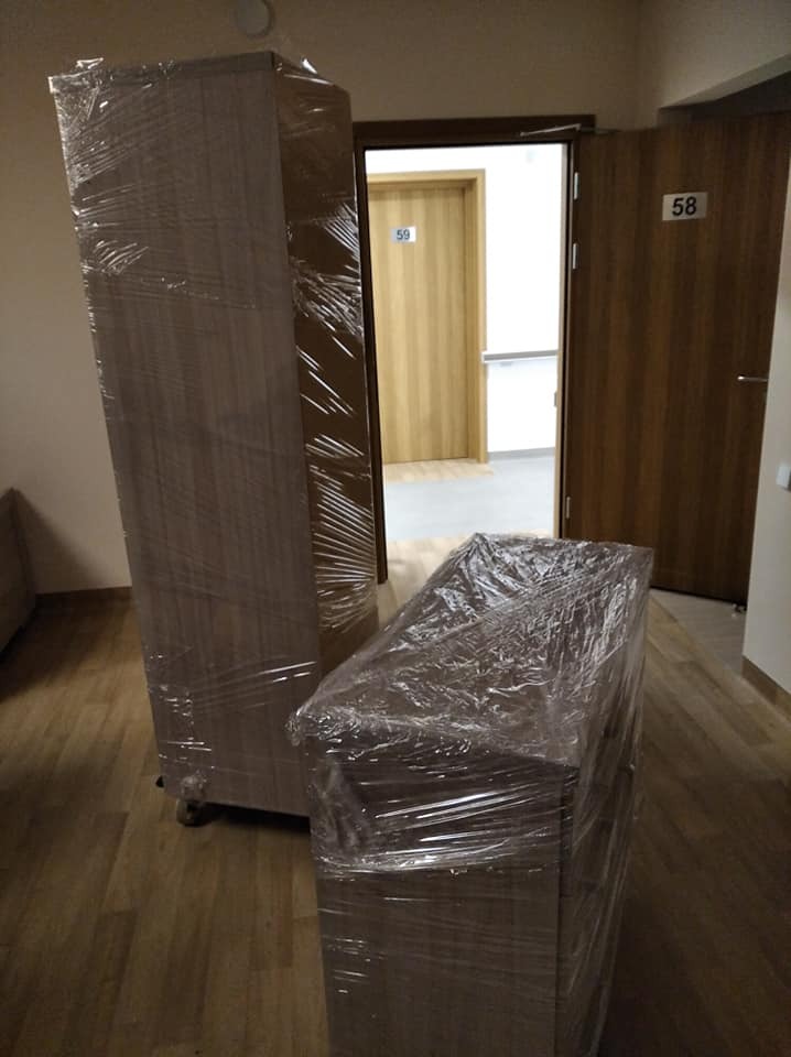 Dwa meble, szafa i komoda, owinięte folią stretch, przygotowane do transportu wewnątrz budynku z numerowanymi drzwiami.