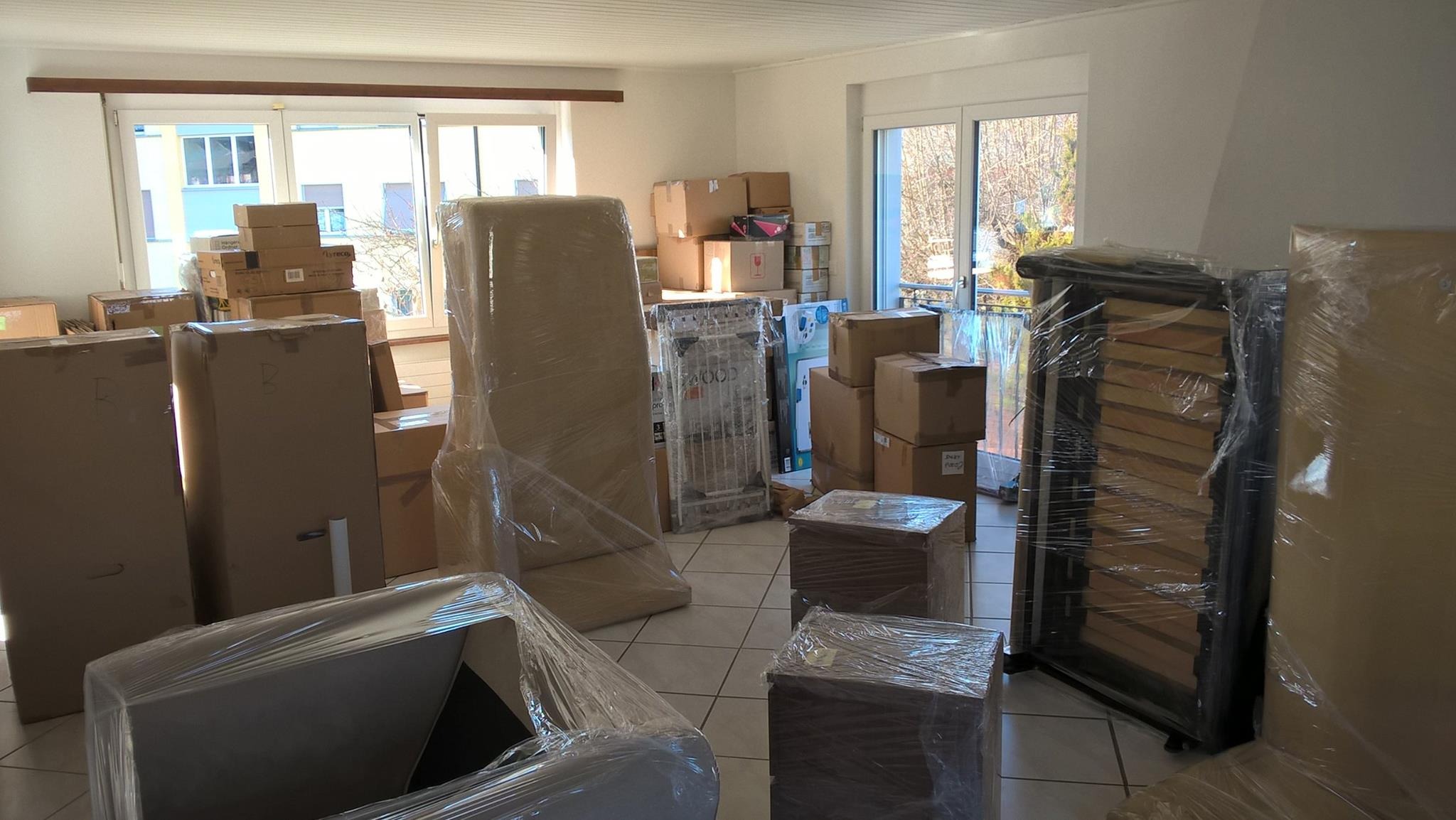 Pokój wypełniony kartonami i meblami owiniętymi folią stretch, przygotowanymi do transportu; widoczne okna balkonowe z widokiem na drzewa.