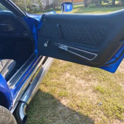 Wnętrze błękitnego klasycznego samochodu z czarną, pikowaną tapicerką drzwi i siedzeń, chromowanymi detalami i widocznymi rurami wydechowymi po bokach.