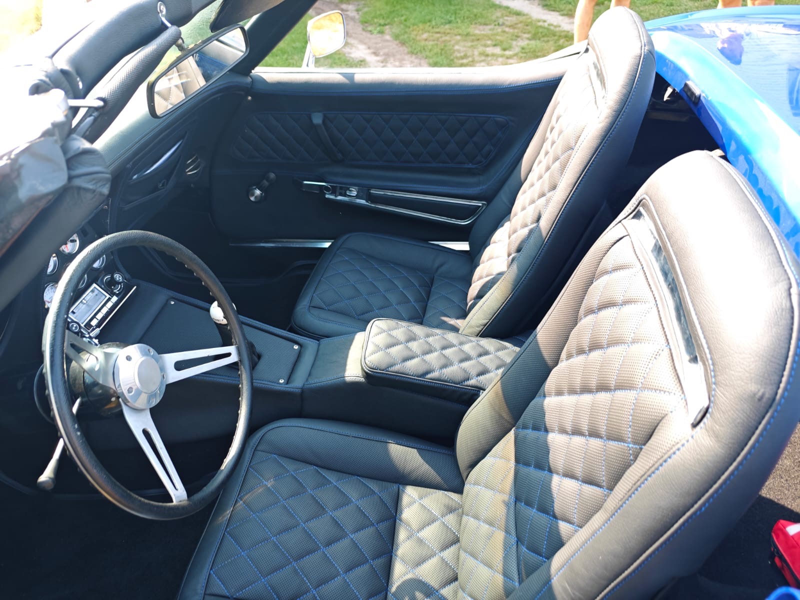 Wnętrze klasycznego kabrioletu z czarną skórzaną tapicerką w romby przeszytą błękitną nicią, widoczna kierownica z trzema ramionami i chromowanymi elementami, oraz radio samochodowe retro.