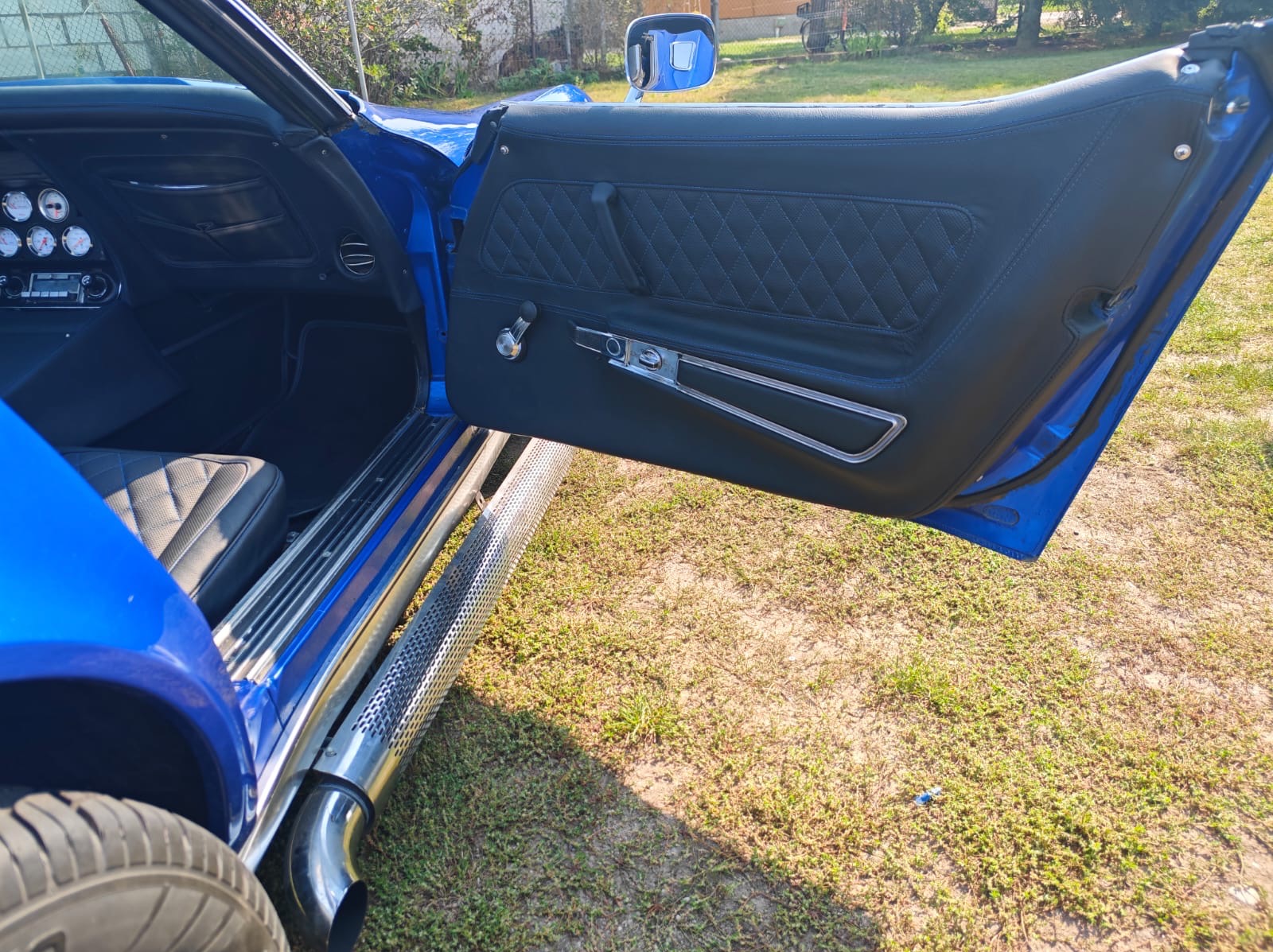 Wnętrze błękitnego klasycznego samochodu z czarną, pikowaną tapicerką drzwi i siedzeń, chromowanymi detalami i widocznymi rurami wydechowymi po bokach.