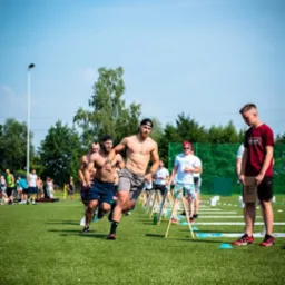 Grupa mężczyzn biegnie przez tor przeszkód na zielonym boisku; widoczny mężczyzna bez koszulki, z brodą i czapką, w szarych spodenkach, prowadzi bieg, a za nim podążają inni uczestnicy; w tle...