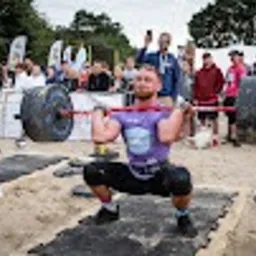 Mężczyzna w fioletowej koszulce wykonuje przysiad z bardzo ciężką sztangą z oponami na piasku podczas zawodów strongman, w tle widoczni kibice i sędziowie.