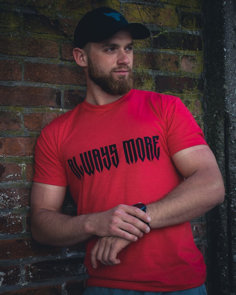 Mężczyzna z brodą w czerwonej koszulce z napisem 'ALWAYS MORE' oparty o ceglaną ścianę, noszący czapkę z daszkiem i sportowy zegarek.