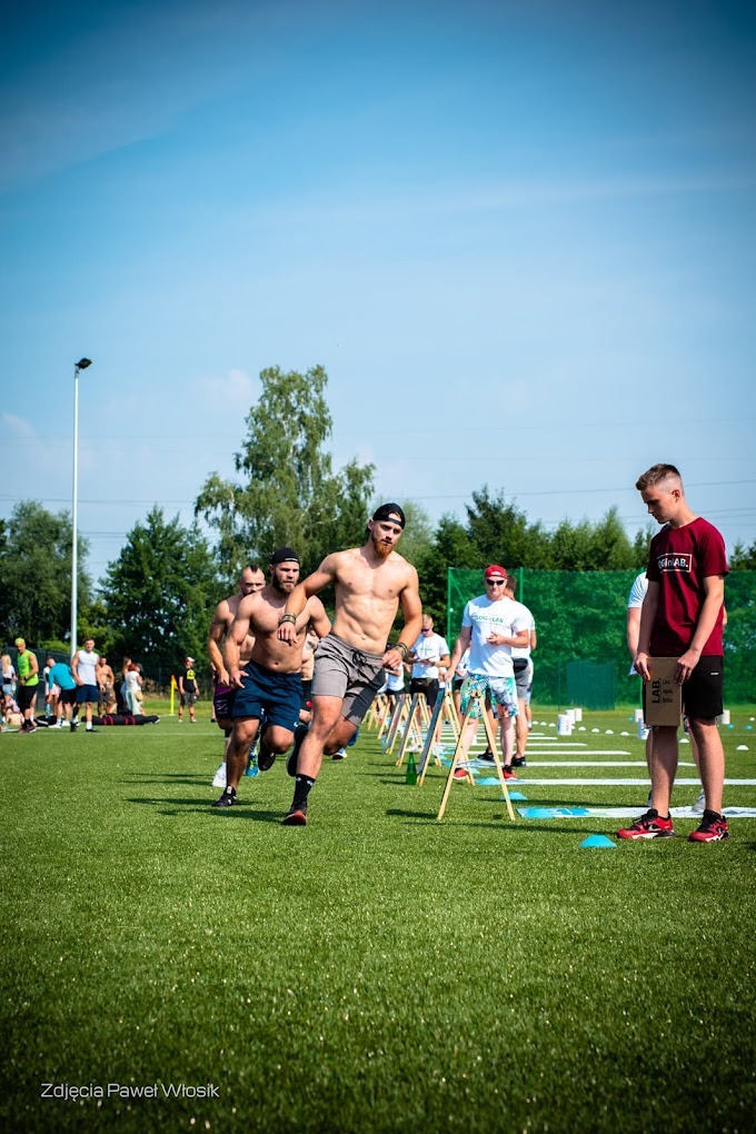 Grupa mężczyzn biegnie przez tor przeszkód na zielonym boisku; widoczny mężczyzna bez koszulki, z brodą i czapką, w szarych spodenkach, prowadzi bieg, a za nim podążają inni uczestnicy; w tle...